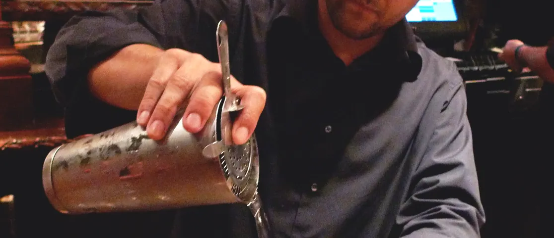 Bartender pouring a craft cocktail at a lively bar counter with glassware in Manhattan